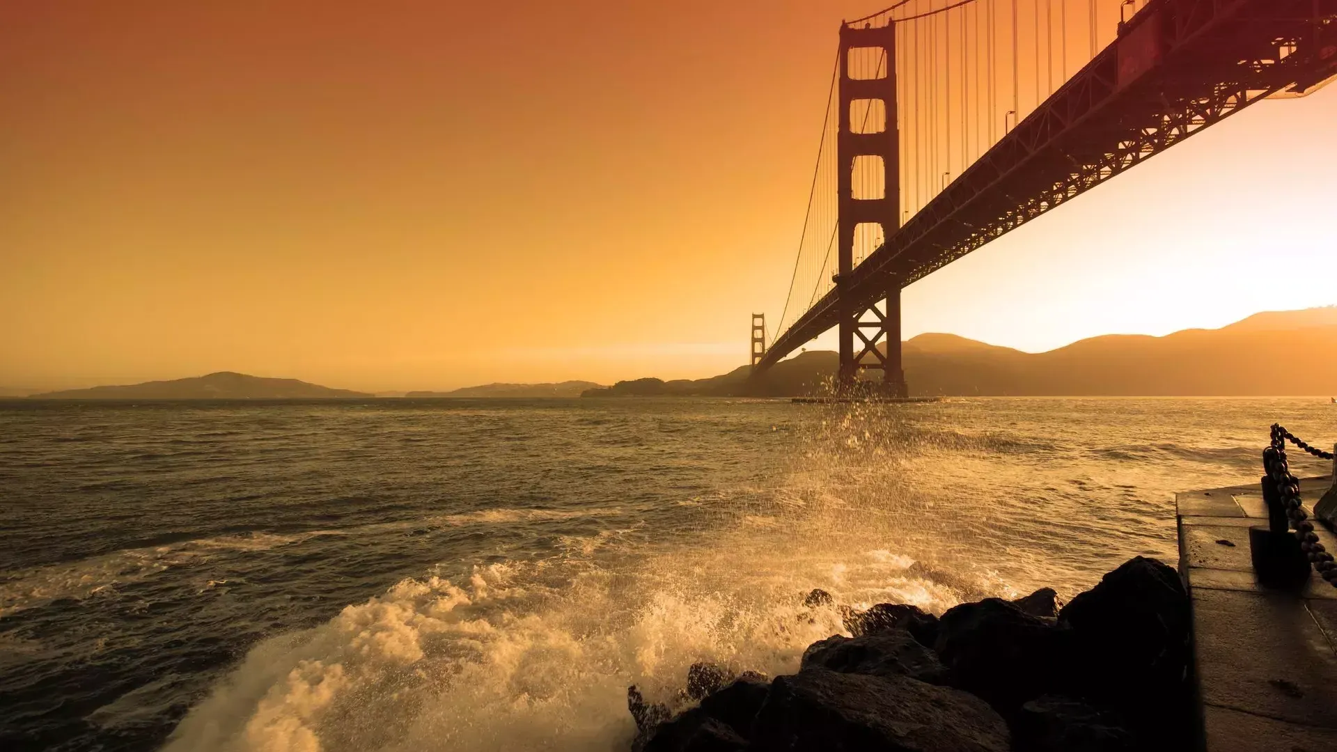 Waves crash near Fort Point beneath the Golden Gate Bridge at sunset.